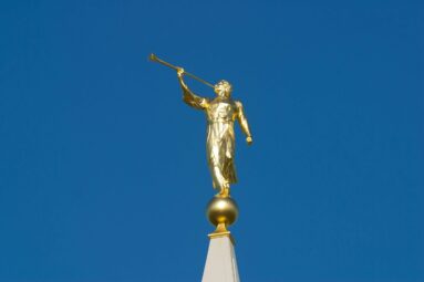 A radiant golden statue of the Angel Moroni, a significant religious figure, stands atop a temple spire against a clear, vibrant blue sky. This iconic symbol represents faith and divine messages, often seen on Latter-day Saint temples worldwide.
