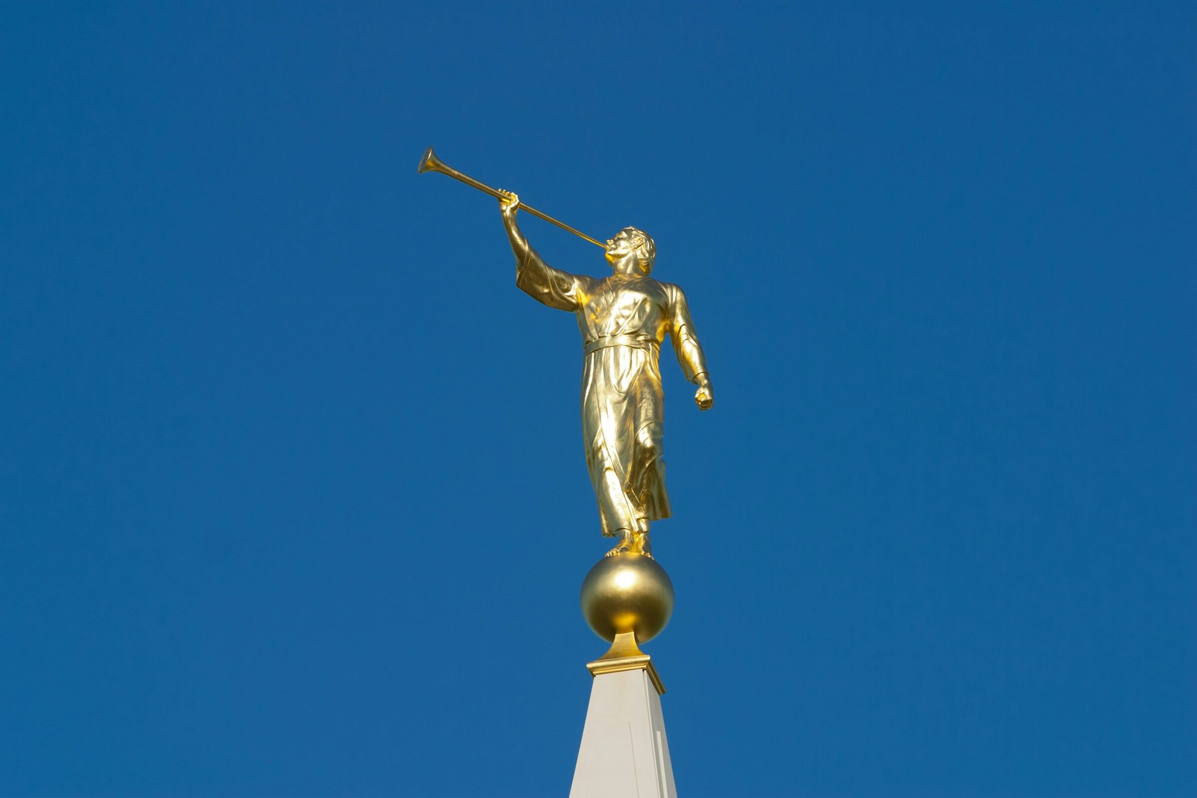 A radiant golden statue of the Angel Moroni, a significant religious figure, stands atop a temple spire against a clear, vibrant blue sky. This iconic symbol represents faith and divine messages, often seen on Latter-day Saint temples worldwide.