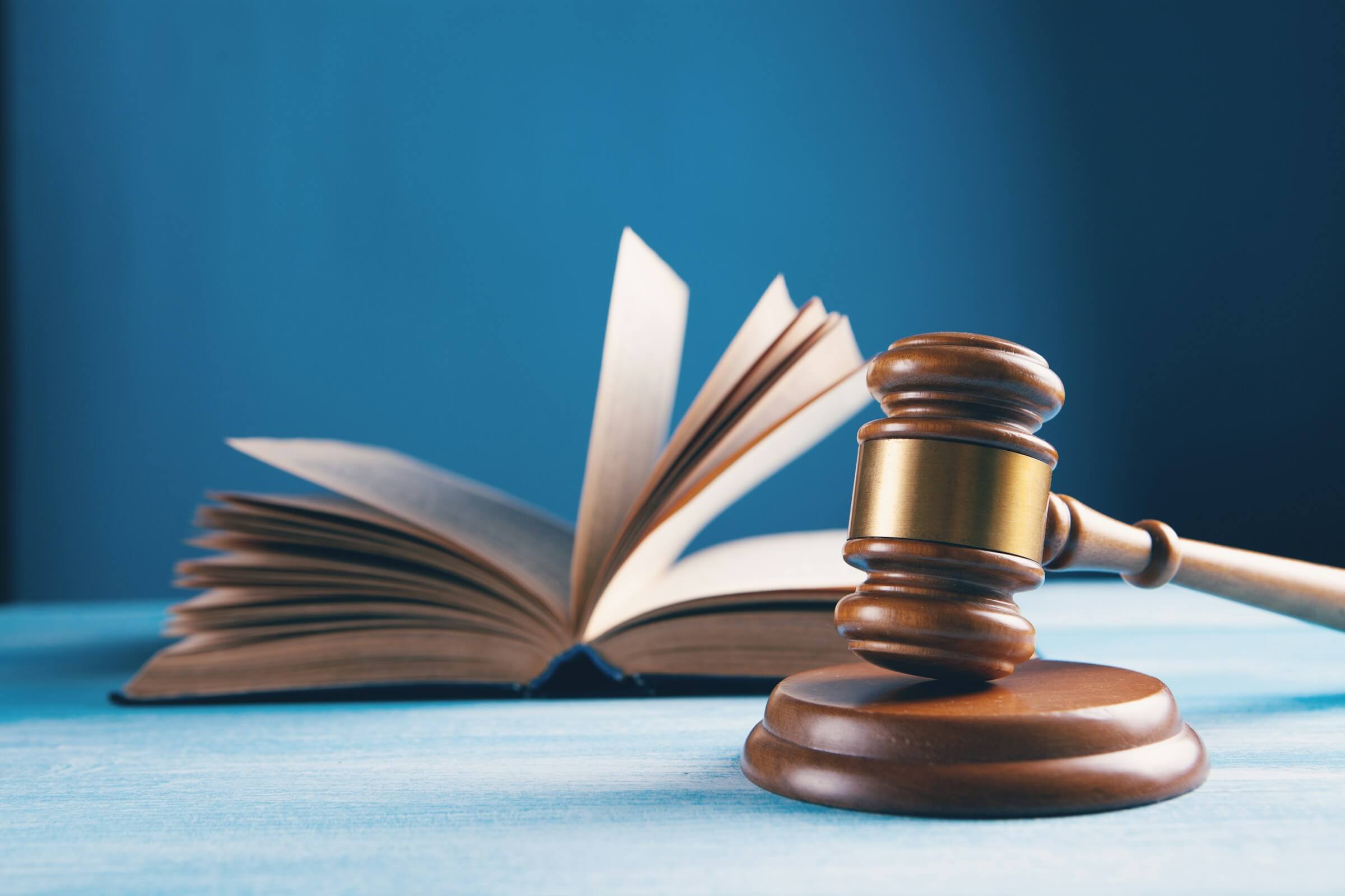 A wooden gavel rests on a sound block in the foreground, with an open book blurred in the background, symbolizing law, justice, and legal education.