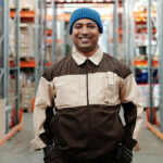 Smiling male warehouse worker in a blue beanie, brown and beige uniform, and gloves, standing confidently in an aisle with shelves of inventory.