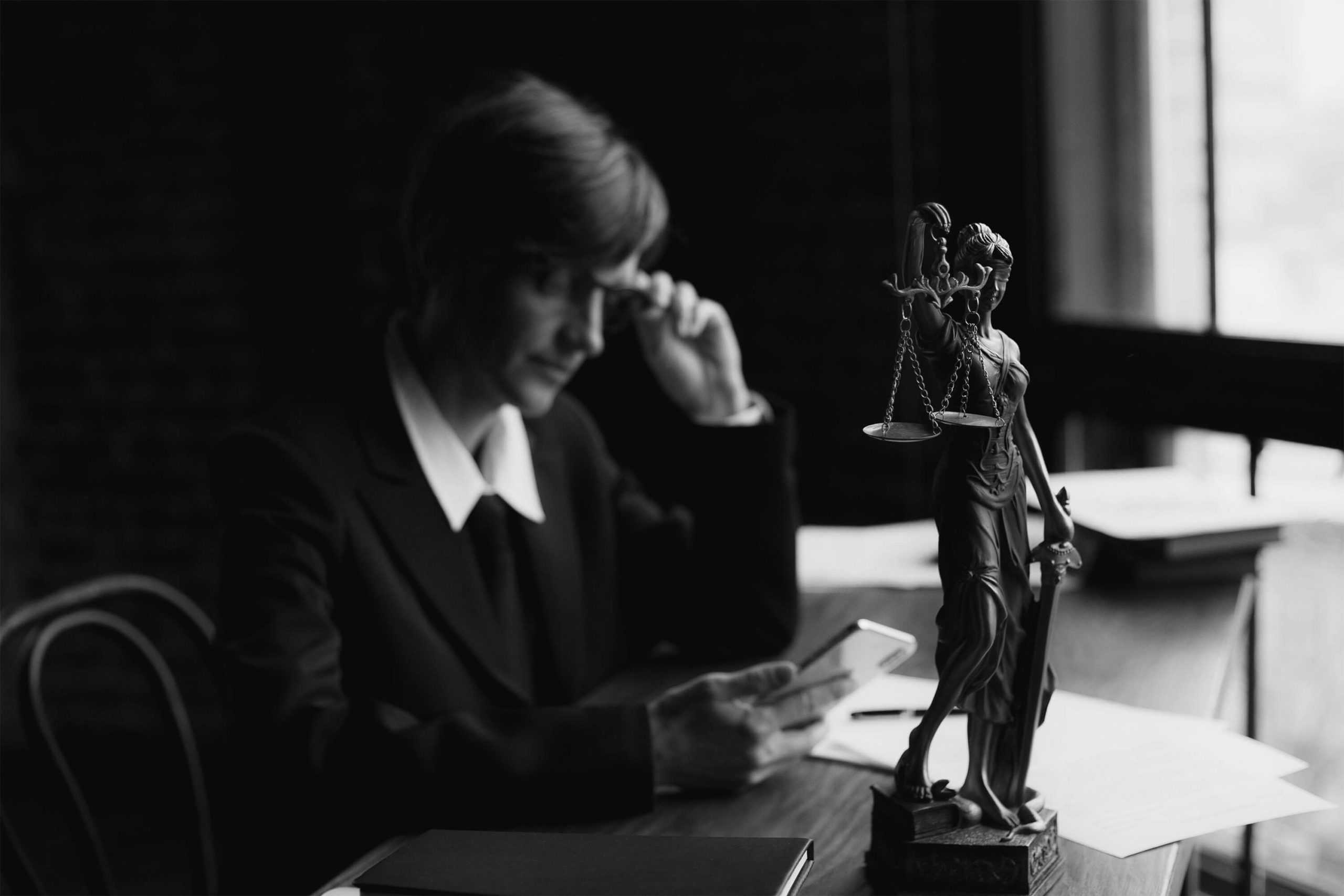 A black and white image of a focused female lawyer in a suit, reviewing legal documents on her phone, with a Lady Justice statue prominently displayed on her desk, symbolizing law, justice, and legal services.